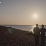 Two people walk side by side along a pebble beach at sunset. The sun hangs low over the sea, reflecting light across the water. Beach huts line the shoreline in the distance on the left, and wooden groynes extend toward the water. The figures appear as dark silhouettes against the bright sky. Visible text reads “HEARTSTOPPER FOREVER” in the top left and a Netflix “N” logo in the top right.